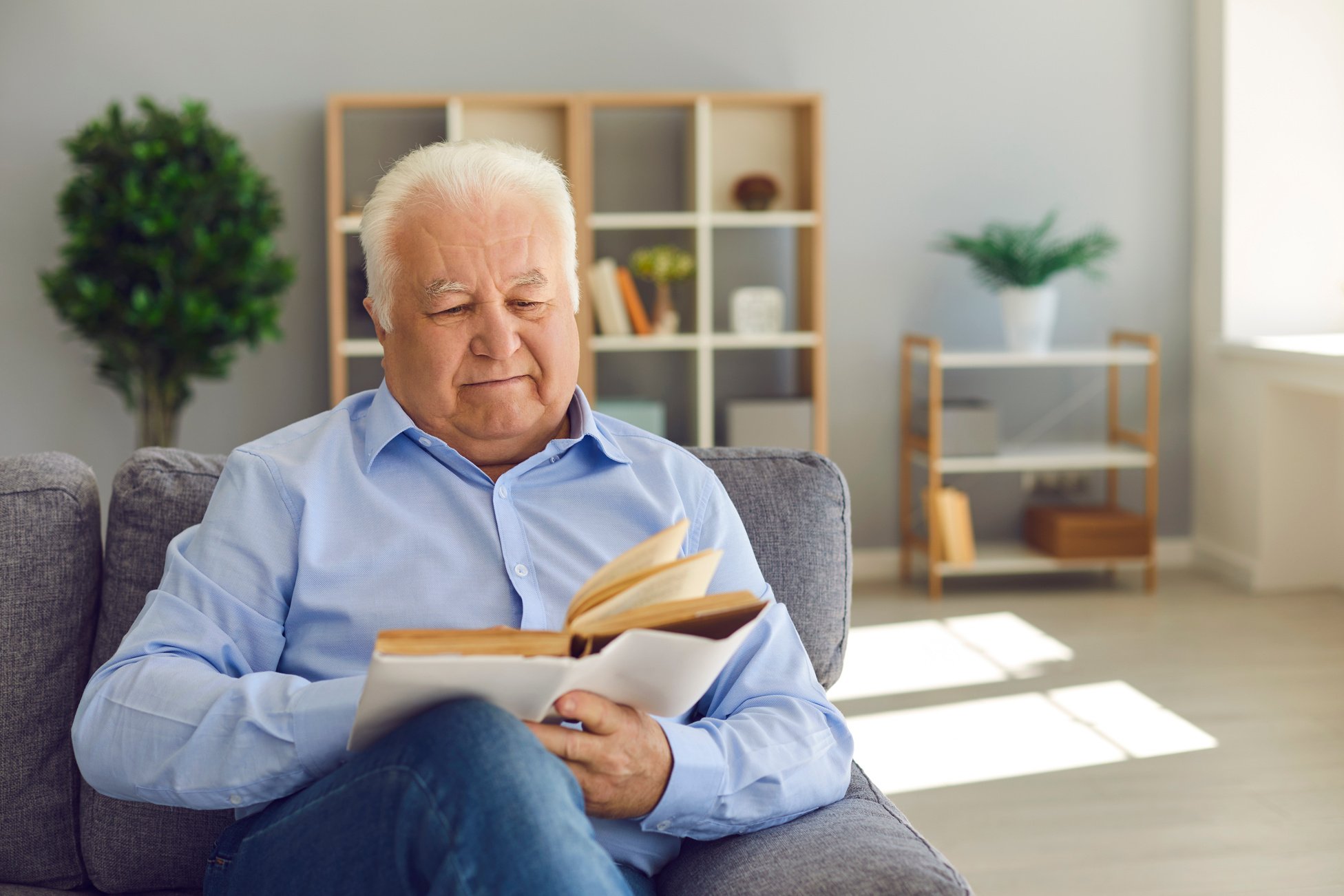 Happy Retired Man Spending Leisure at Home and Reading Old Book Sitting on Couch in the Living Room